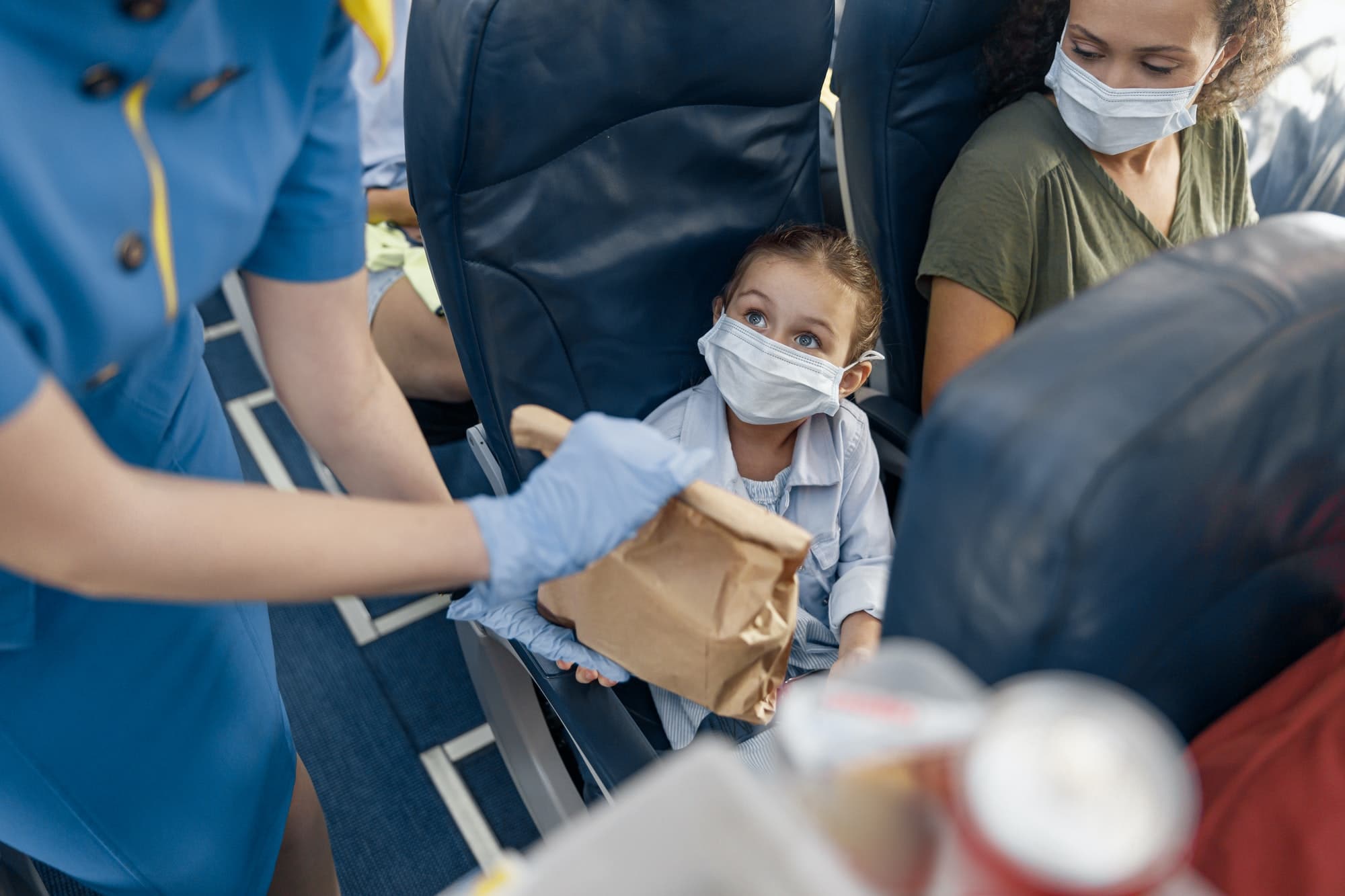 Cute girl in protective face mask looking at flight attendant serving lunch to little passenger